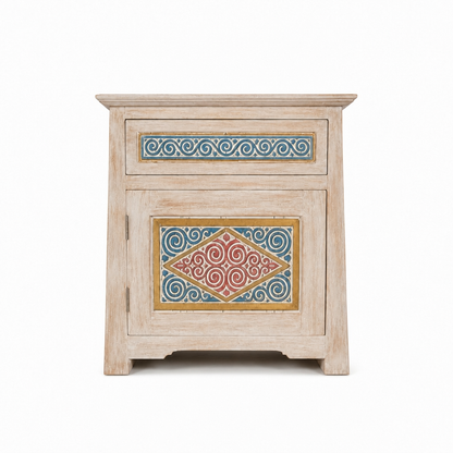 A small rustic wooden nightstand with a distressed light beige finish, featuring a single drawer and a lower cabinet door, both decorated with colorful hand-painted geometric and scroll patterns in blue, red, and gold.